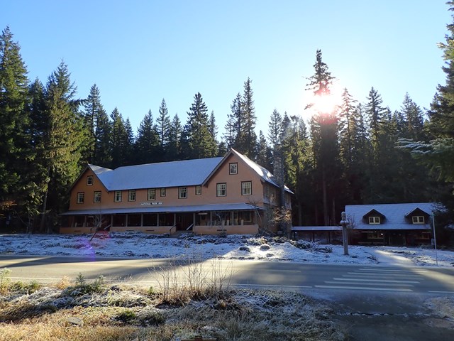 A two-story wood building with a small building next door, dusted by snow and surrounded by forest.