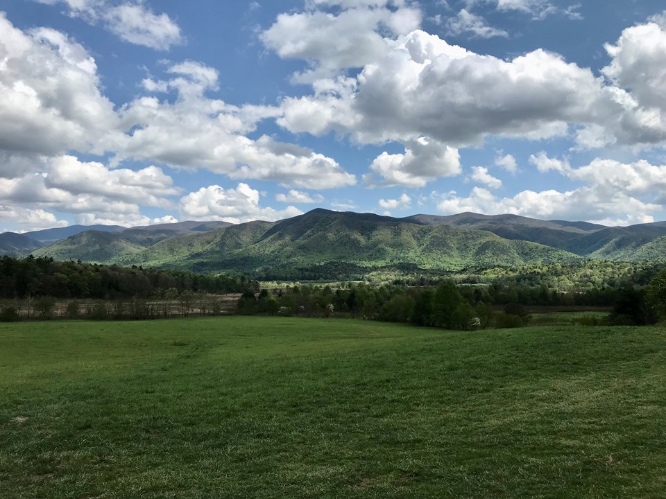 A grassy valley surrounded by distant mountains with blue sky and clouds above.