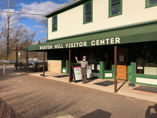 Female ranger wearing a mask and uniform waves hello on porch of a cream building with green awning.