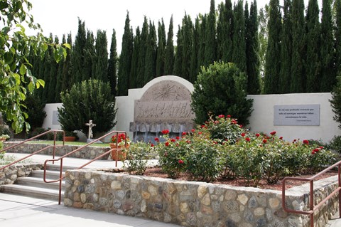 A bed of roses in front of a large wall fountain and grassy area around a grave marker