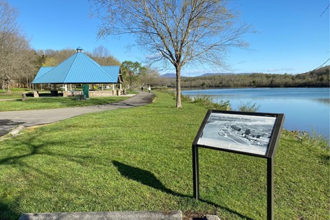 An historical marker on the bank of a calm river with a paved trail and pavilion in the background.