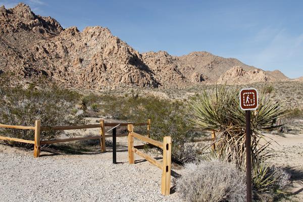 A dirt trail starting between wooden fences with rocky mountains in the distance.