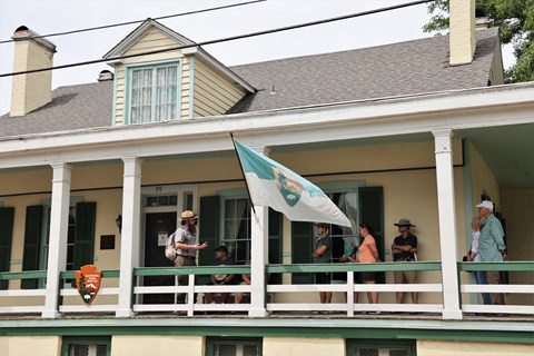 Park ranger talking under a covered porch of a cream house to several people.