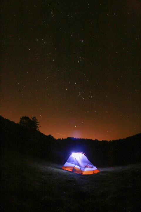 An illuminated two-person tent under a starry sky with orange light pollution on the horizon.
