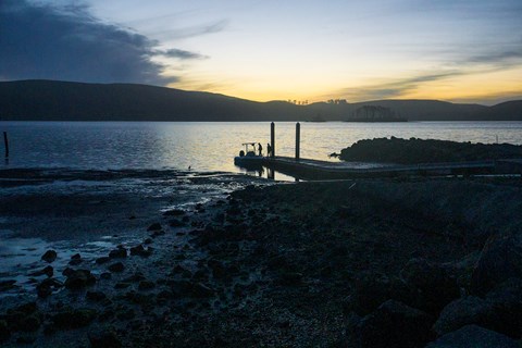 Two persons stand on a floating dock adjacent to their motor boat shortly after sunset.