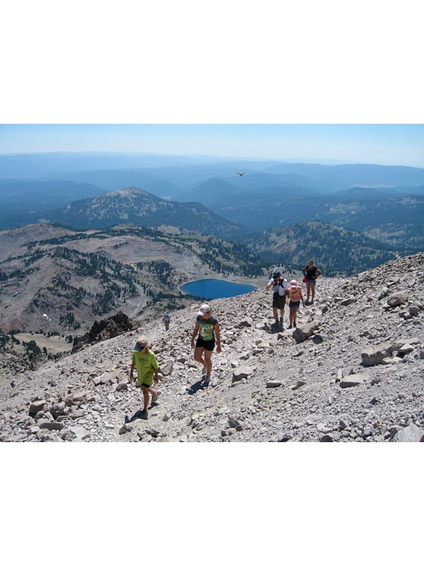 A family hikes up a rocky a trail above a bright blue alpine lake and distance volcanic peaks.