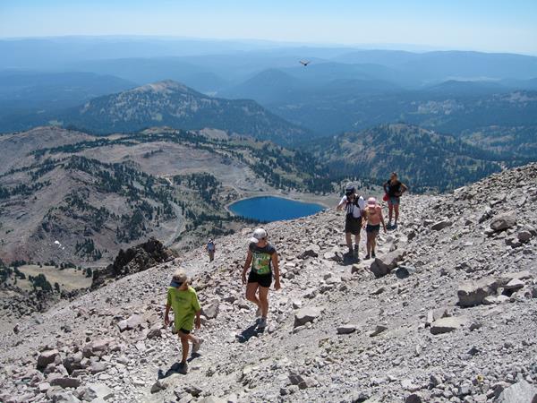 A family hikes up a rocky a trail above a bright blue alpine lake and distance volcanic peaks.
