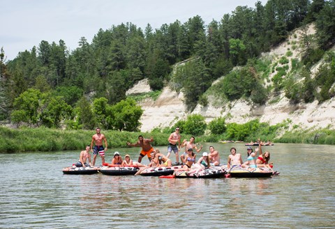 a collection of 7 tubes on the river, with 13 people floating on them; they are tied together.