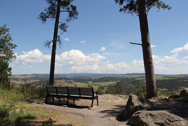 A bench on a gravel trail between two ponderosa pines, overlooking a forested valley and distant hills