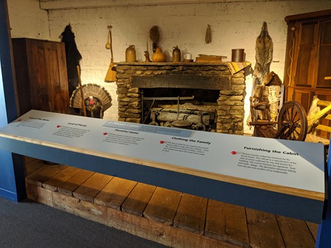 Exhibit of inside a cabin with hearthstones, cabinets, spinning wheel, furs and more.