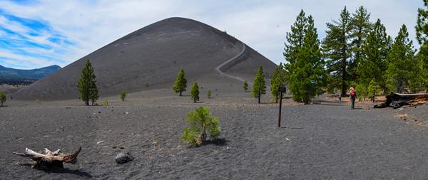 A panoramic view of a cinder cone volcano with sparse conifers at its edges. Two hikers walk on a su