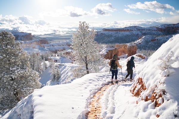 Two hikers walk on a snow covered trail with mountains in the background.