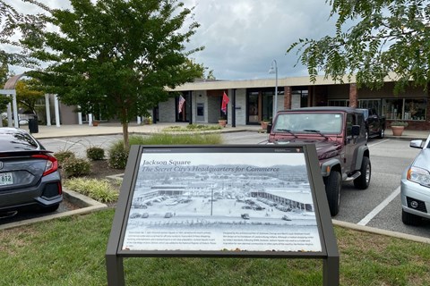 A strip mall with cars parked and historical marker in the foreground.