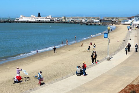 various people and children sit or play in the sand on the beach near Hyde Street Pier.