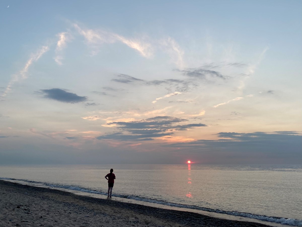 Lake Michigan beach at sunset with a hazy red sun on the horizon under a blue, cloudy sky.
