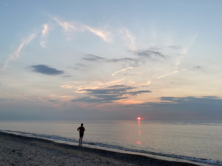 Lake Michigan beach at sunset with a hazy red sun on the horizon under a blue, cloudy sky.