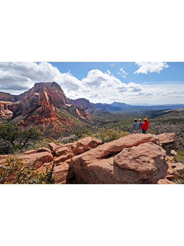 Red sandstone rocks with three hikers standing on them looking out over a valley. Blue, cloudy sky.