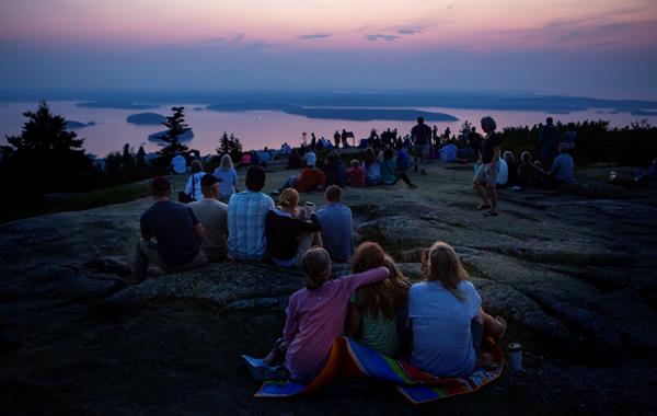 Groups of people sit on a granite outcropping, looking out at a view of sunrise over ocean.