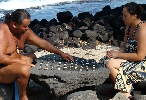 A man and and woman in traditional clothing sit at a kōnane board. The man reaches for a game piece.