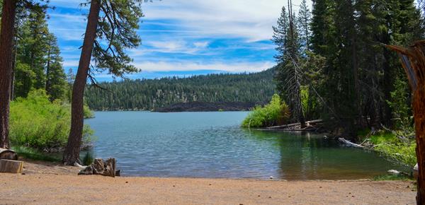 A gravel boat launch area at the edge of a large lake lined by conifers and bushes.