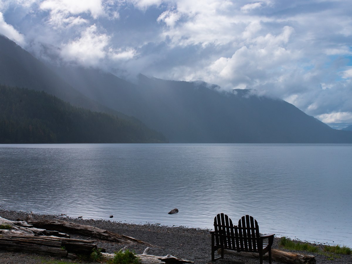 A wooden love seat next to a peaceful lake.