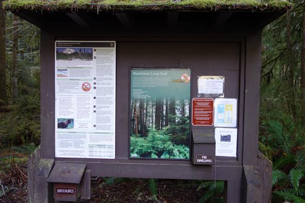 A vertical exhibit panel attached to a information board covered by a shallow roof covered in moss.