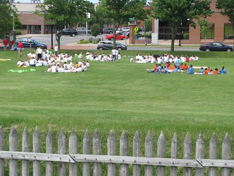 A large group of children sit on blankets scattered across the lawn.