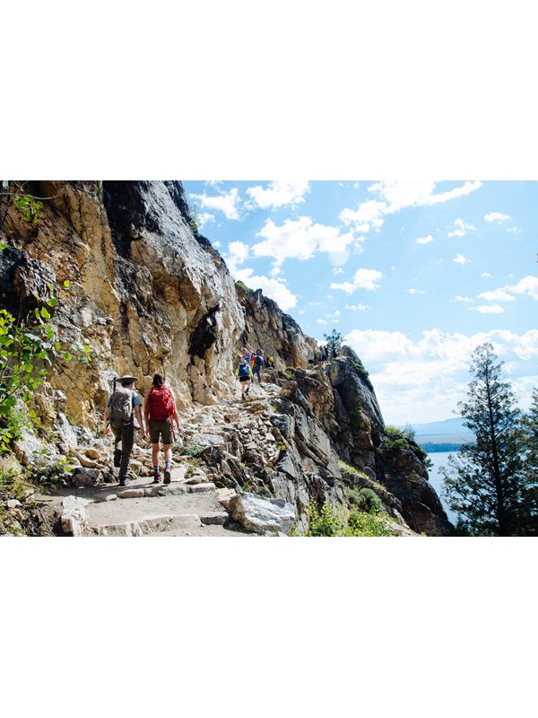 Hikers walk up a rocky trail with a cliff on the right.