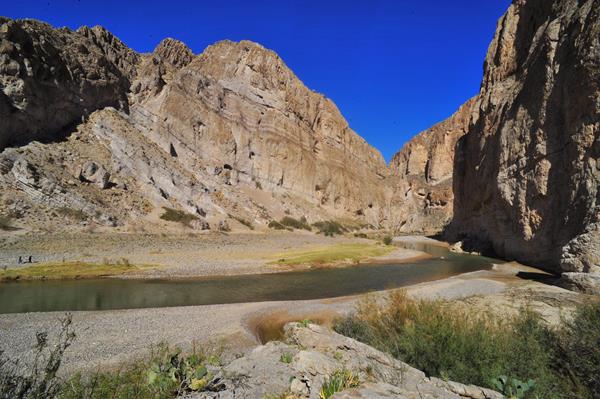 A river flows through high walls of limestone, while hikers walk along the sandy banks.