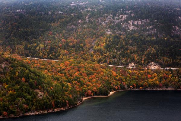 A road goes through brightly colored leaves around a mountain above a lake.