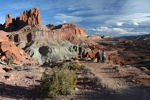 Two hikers stand at the edge of a cliff, looking at a panoramic view of colorful rock cliffs.