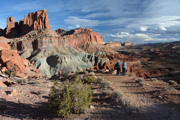 Two hikers stand at the edge of a cliff, looking at a panoramic view of colorful rock cliffs.