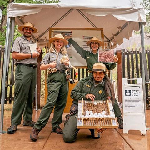 Four NPS rangers stand under a shade structure holding various props.