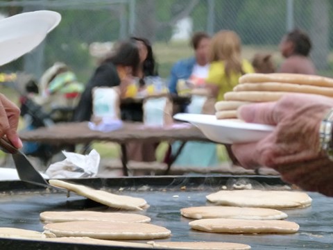 Close-up of pancakes on a griddle being served. People eat at picnic tables in the background.