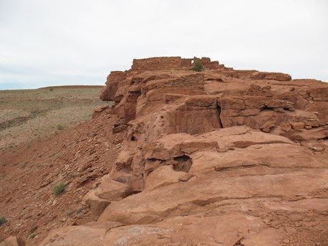 A partial sandstone pueblo on top of a rock outcrop with grassy hills in the distance.