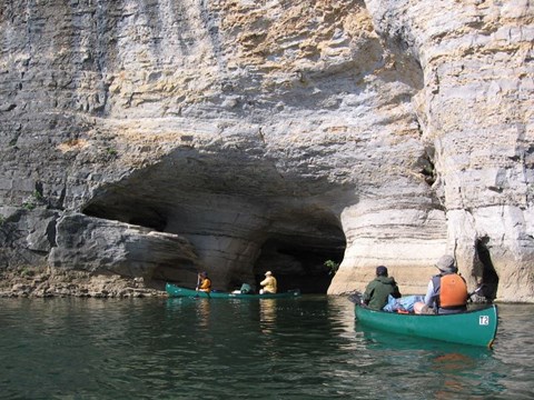 green canoes on river in front of grey cliff with large hole at river level