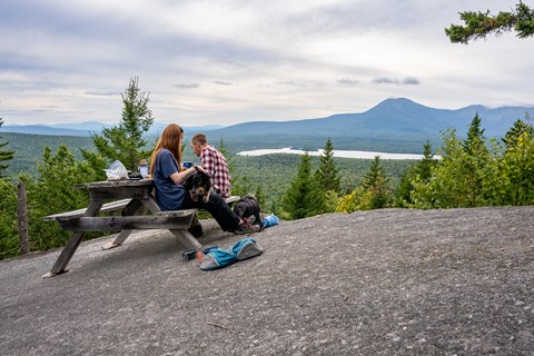 Two hikers and their dog on a leash have a picnic with a view on top of Barnard Mountain.