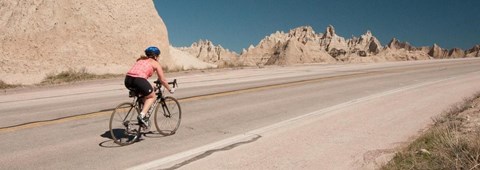 a woman bikes up a paved road with jagged badlands spires in the background