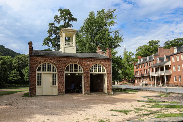 Small one-story red brick building with three arched doorways and a white cupola, standing on a gravel lot beside brick row houses