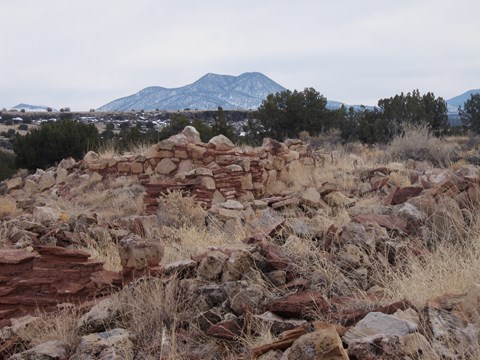 The remnants of a tan stone pueblo structure surrounded by yellow grasses and juniper trees.