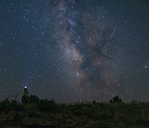 A hiker with a headlamp looks up at the star-filled night sky. The Milky Way Galaxy creates a band o