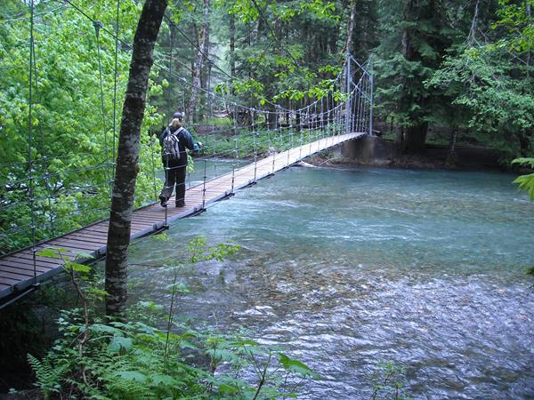 A hiker walks across a suspension bridge in the forest above bright blue water.