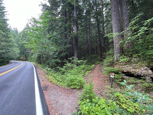 To the east side of the two-lane paved highway is a narrow, dirt trail surrounded by trees.