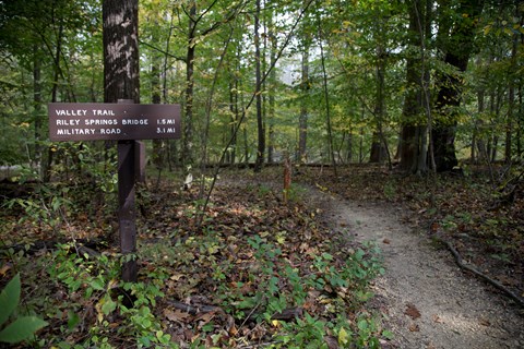 A brown and white sign with a trail in the background.
