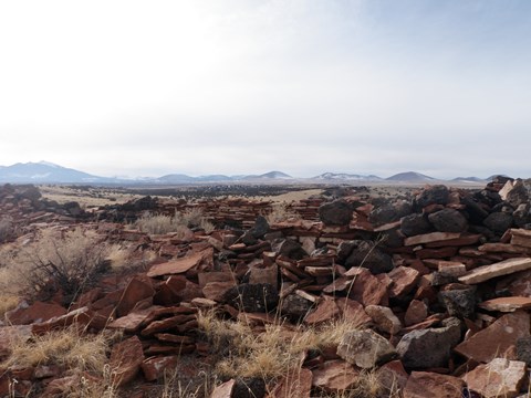 The remnants of a sandstone and basalt pueblo structure with volcanic hills in the background.