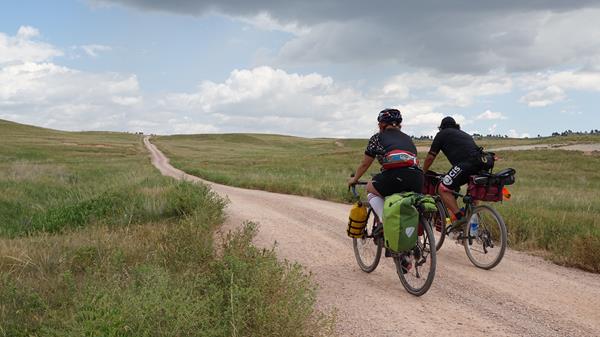 two people in biking gear riding bikes on a dirt road in the open prairie