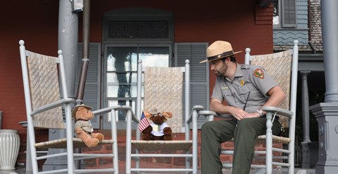 A Park Ranger sits in a rocking chair next to a stuffed moose toy and a stuffed teddy bear.