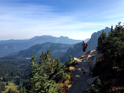A backpacker on a rocky trail pauses to overlook a forested valley and mountain ridges.