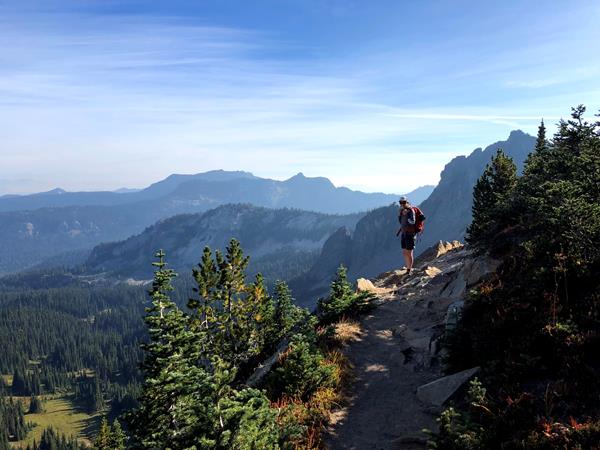 A backpacker on a rocky trail pauses to overlook a forested valley and mountain ridges.