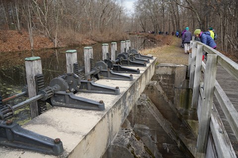 Group of people walking down path with industrial mechanisms visible on the left side of image